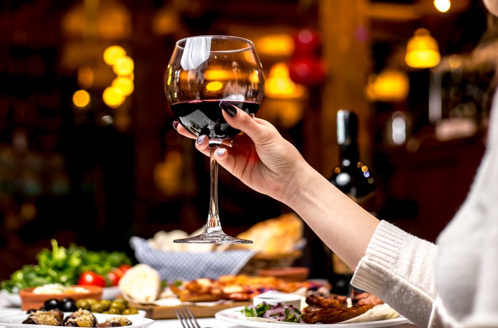 Mano de mujer sostiene una copa de vino tinto monastrell sobre una mesa con comida